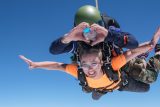 A tandem skydiving pair is free-falling against a clear blue sky. The instructor is wearing a helmet and goggles, forming a heart shape with his hands, while the excited woman in an orange shirt spreads her arms wide.