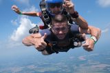 Two people tandem skydiving, with one giving thumbs up. They are free-falling through clear skies, with a landscape below. Both wear helmets and goggles, with the instructor guiding from behind.