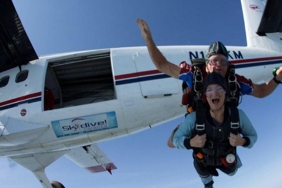 Two people are tandem skydiving, one instructor and one participant. They are exiting a white airplane mid-air, with the open plane door visible. The sky is clear and blue, and both are wearing parachute harnesses. The participant looks excited.