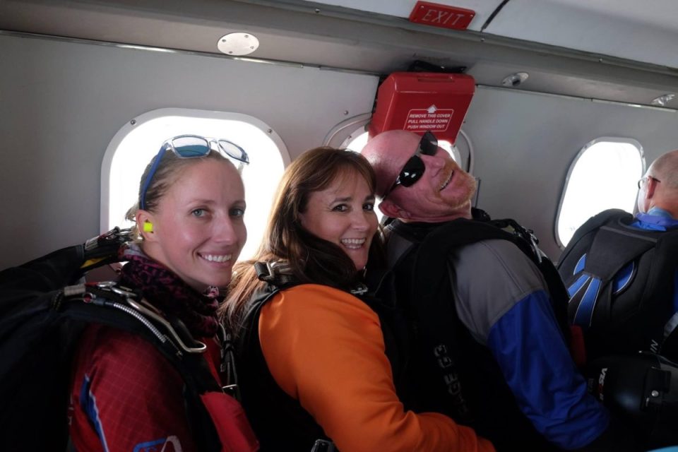 Three people smile inside a plane, wearing skydiving gear. Two women and one man, all with parachute packs, are seated side by side near a small window. The man wears sunglasses, and the woman in the middle wears an orange jacket.