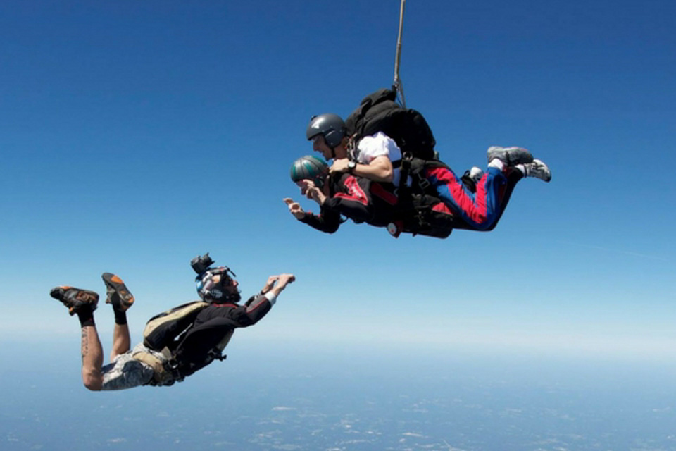 Two people skydive while another person holds and photographs a bowling ball in mid-air. They are all wearing helmets and skydiving gear against a clear blue sky backdrop.