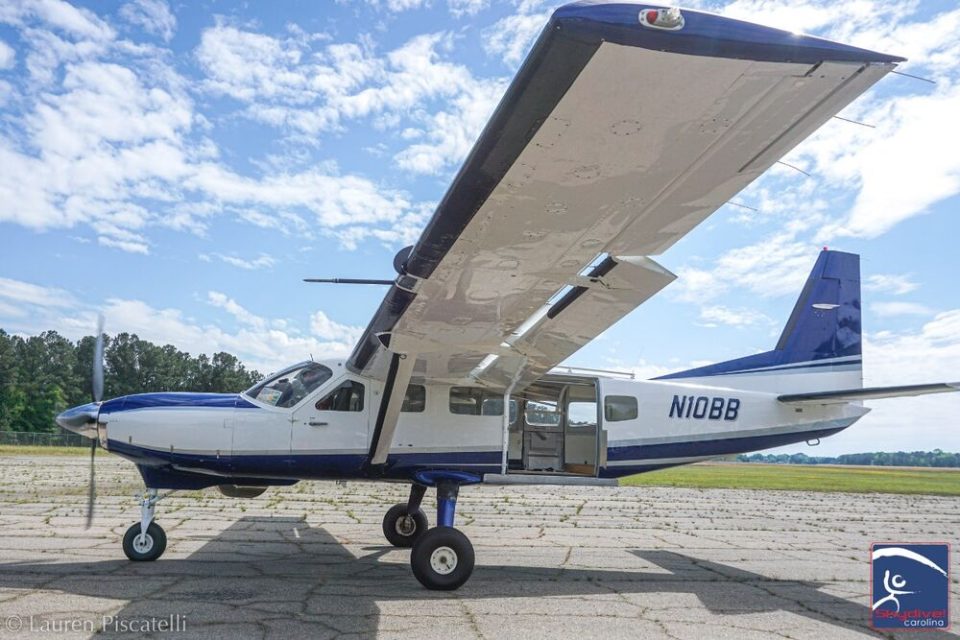 A small blue and white aircraft is parked on a runway under a partly cloudy sky. The plane's door is open, and its distinctive high wings are prominent. Trees and grass line the background.