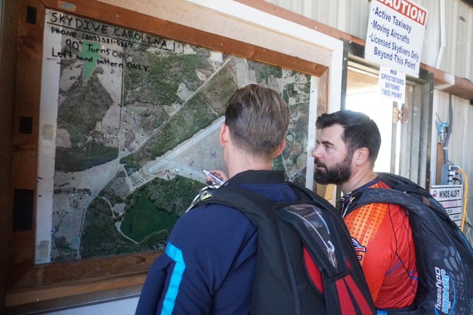 Two people wearing parachute harnesses examine a large aerial map posted on a wall. The map includes flying patterns and safety zones, and the location is identified as Skydive Carolina.