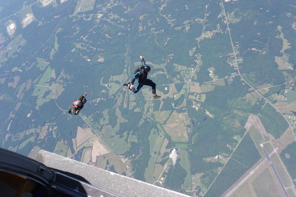 Two people skydiving high above a patchwork of green and brown fields. The open airplane door is visible in the foreground, and the divers are freefalling with parachutes not yet deployed. The landscape features roads, forests, and farmland.