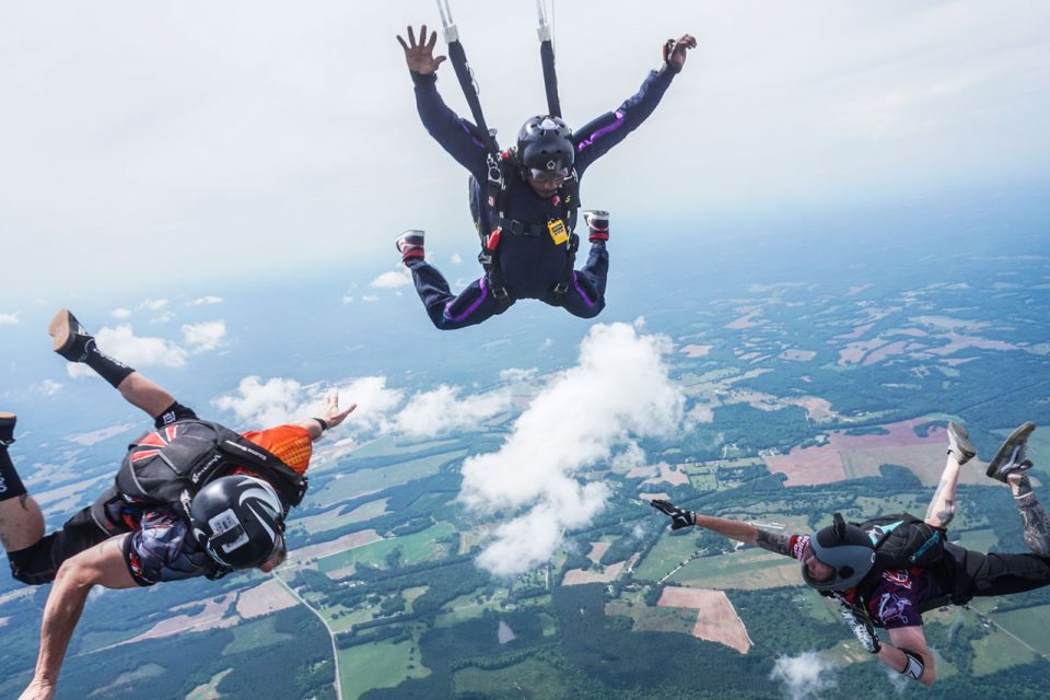 Three skydivers in mid-air, wearing parachute gear, performing acrobatic maneuvers against a backdrop of fields and forests far below. The sky is clear with a few scattered clouds.