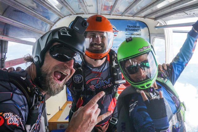 Three skydivers in helmets and gear are excitedly posing inside an aircraft. They are making hand gestures and smiling widely. Outside, bright clouds are visible, suggesting they're ready for a jump.