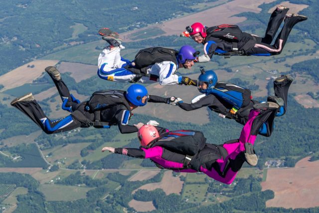 Six skydivers in colorful suits and helmets form a circle while free-falling over a rural landscape with fields and forests. They hold hands and maintain a synchronized formation in mid-air.