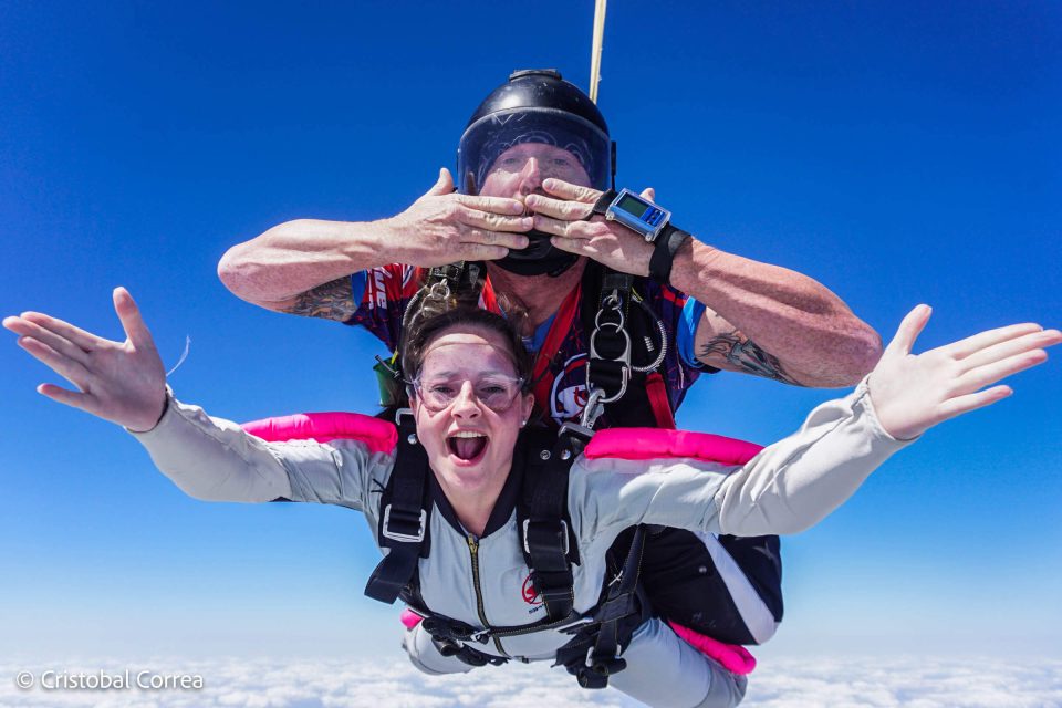 Two people skydiving in tandem, smiling against a clear blue sky. The instructor, behind, makes a playful gesture with his hands. Both are wearing skydiving gear and goggles. Bright, sunny day above the clouds.