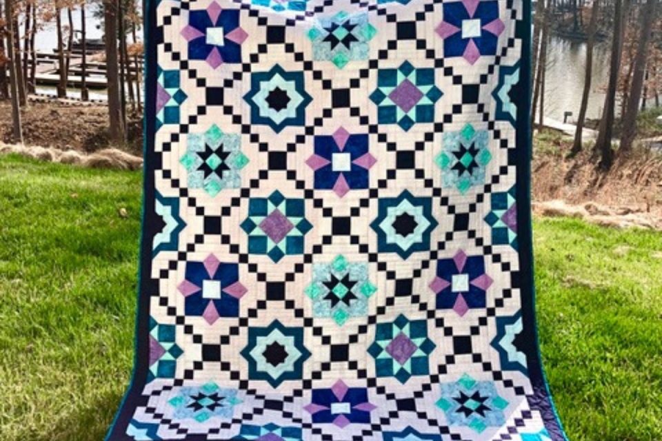 A colorful quilt displayed outdoors on grass, featuring a geometric pattern with star and square motifs in shades of blue, purple, turquoise, and white. In the background, there's a pond surrounded by trees.