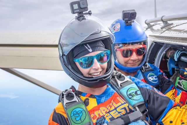 Two skydivers in colorful jumpsuits and helmets with mounted cameras prepare to jump from an airplane. Both are smiling and wearing sunglasses. The sky is visible in the background.