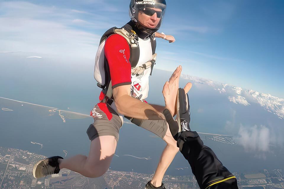 Two skydivers performing a high-five in mid-air against a backdrop of a coastal landscape. One wears a red and gray outfit, the other in dark gear, while the ocean and clouds add to the thrilling scene below.