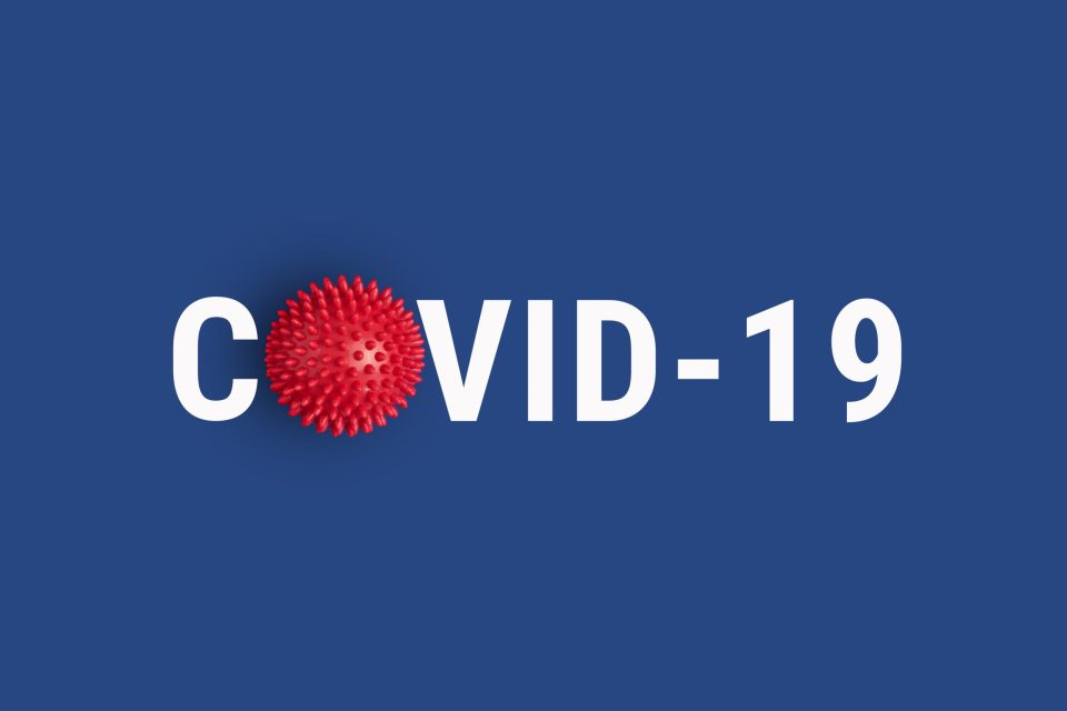 The image shows the text "COVID-19" against a blue background. The "O" is replaced by a red spiky coronavirus illustration.