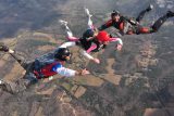 Four people are skydiving in formation over a landscape of fields and trees. They are in freefall, wearing helmets and jumpsuits, with one person in a red helmet leading the group. The ground is far below them.