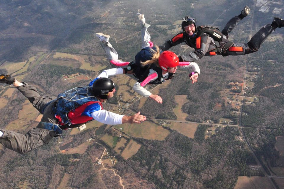 Four people are skydiving in formation over a landscape of fields and trees. They are in freefall, wearing helmets and jumpsuits, with one person in a red helmet leading the group. The ground is far below them.