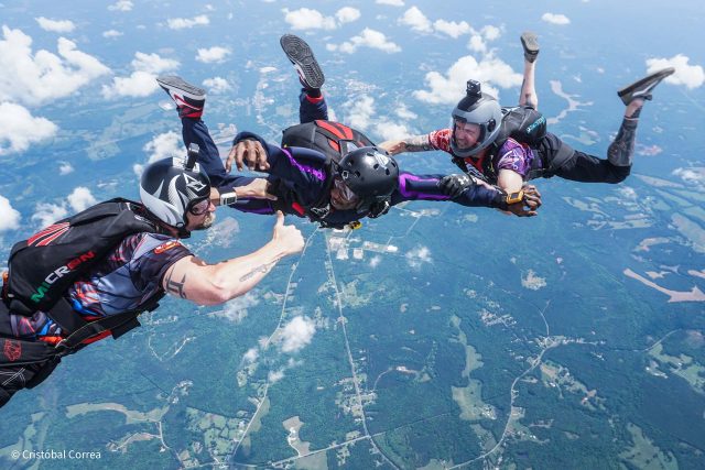 Three skydivers in mid-air form a close formation above a landscape of forests and fields. All are in freefall with visible parachute packs, wearing helmets and gloves, capturing the thrill and teamwork of skydiving.