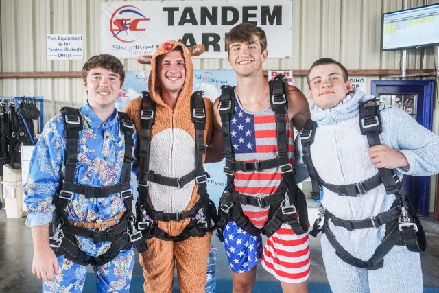 Four people in colorful outfits and parachute harnesses pose indoors, smiling. They stand in front of a sign that reads "Tandem" and "Skydive Carolina." Two wear pajamas, one a reindeer suit, and another an American flag outfit.