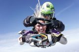 Two skydivers in freefall make triangular hand gestures while smiling. The instructor wears a green helmet and sunglasses. Clear blue sky in the background adds to the exhilarating atmosphere.