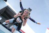 A woman in a tandem skydive with an instructor, both wearing harnesses, exits an airplane. She smiles and raises her hand in a playful gesture, wearing sunglasses and casual clothes. The background is a clear blue sky.