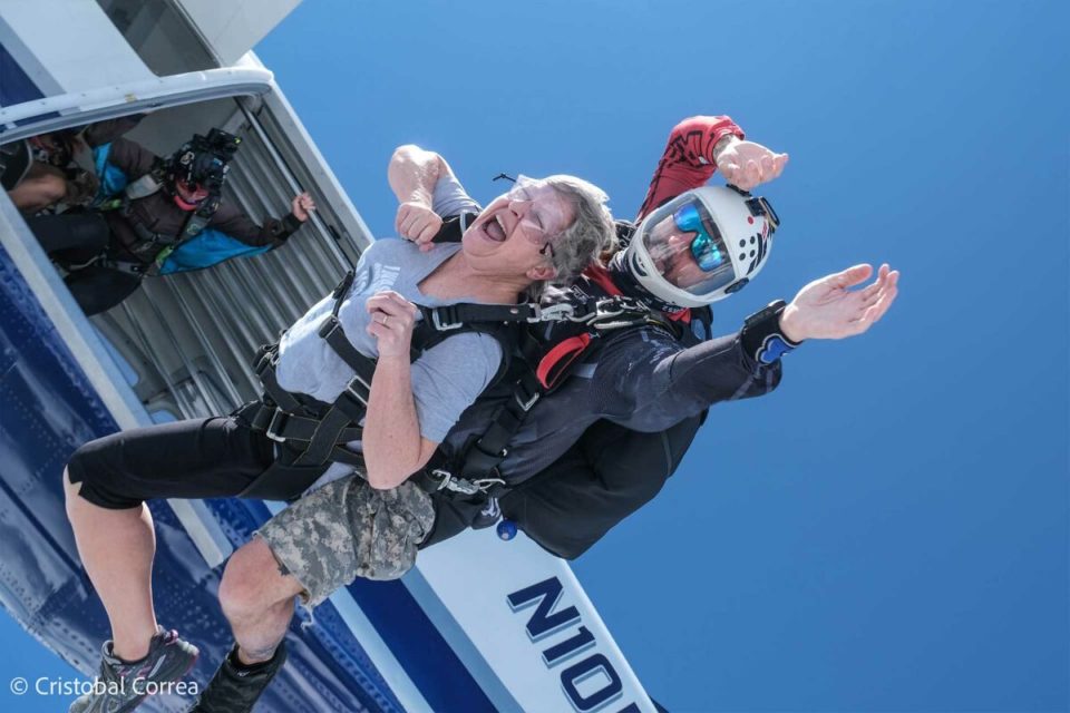 An older woman and her tandem skydiving instructor jump out of a plane, both wearing parachute gear. The woman looks thrilled and nervous, while the instructor controls their descent. The sky is clear and blue. A photographer captures the moment from the plane.