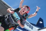 Two people tandem skydiving, with the person in front wearing goggles and an expressive look. They are exiting a plane mid-air, with one person's arms raised in excitement. The sky is clear and blue.