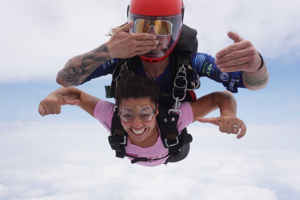 A woman is tandem skydiving with an instructor. She is smiling widely, wearing a pink shirt and goggles. The instructor is above her, wearing a red helmet and a blue suit. They are surrounded by a clear sky and clouds.