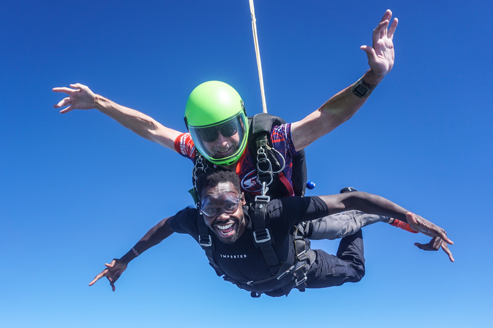 Two people are tandem skydiving against a clear blue sky. The front person is smiling widely, wearing sunglasses and a black jumpsuit. The instructor behind wears a green helmet and colorful gear, both with arms extended outward.