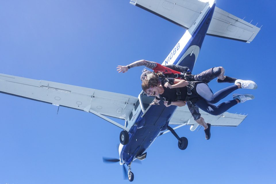 Two people are tandem skydiving from a small aircraft. They are in free fall against a clear blue sky. The plane is shown above them with one person wearing a harness attached to the instructor.