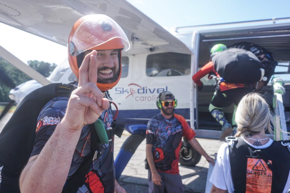 A group of skydivers prepares to board a plane. One person in the foreground smiles and flashes a peace sign, wearing a helmet and gear. Others are gathering near the plane. The background shows blue skies and the plane with "Skydive Carolina" written on it.