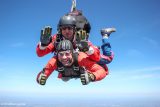 Two people skydiving; the front person smiles and waves at the camera while wearing a helmet and goggles. The instructor behind them is also wearing safety gear. They are surrounded by a clear blue sky.