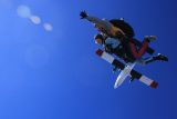 Two people skydiving in tandem against a bright blue sky, with a small airplane in the background. They are wearing parachute gear and appear to be enjoying the experience, with one person raising an arm in excitement.