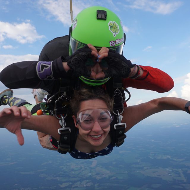 Two people tandem skydiving against a backdrop of blue sky and distant clouds. The person in the front is smiling and wearing goggles, while the person behind is making a hand gesture. Both are wearing helmets and gear.