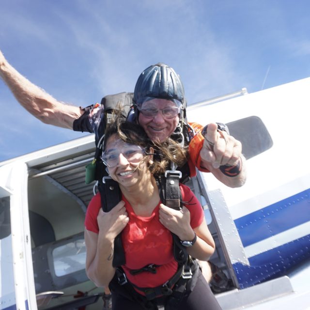 Two people tandem skydiving, wearing goggles and harnesses, just exited a plane. The instructor, behind, smiles and gestures to the camera, while the skydiver in front beams with excitement. The plane and blue sky are visible in the background.