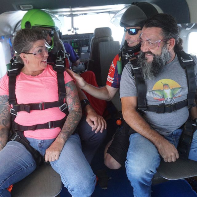 A group of people in a plane, wearing skydiving gear. A woman in a pink shirt and a man with a beard, both with parachute harnesses, sit on a bench. Two men guide them, one checking equipment. The cabin is cramped with skydiving gear visible.