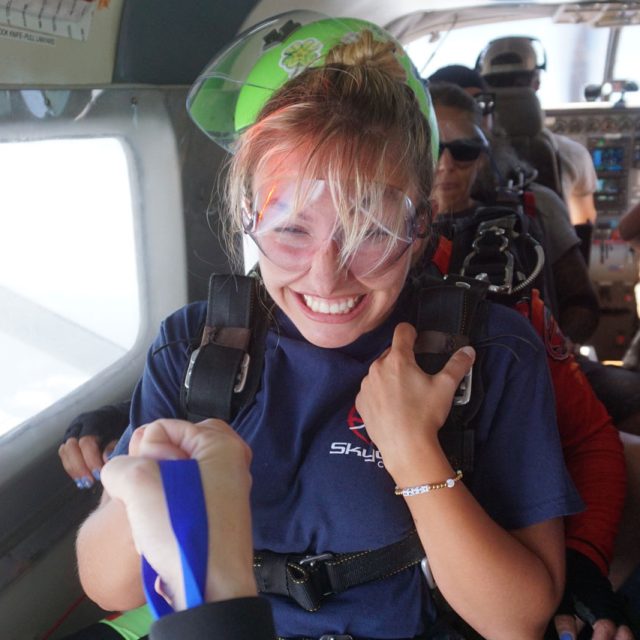 A smiling woman wearing a parachute harness and goggles sits inside a plane, preparing for a skydive. She holds onto a wrist strap and appears excited. Other skydivers are seated behind her.