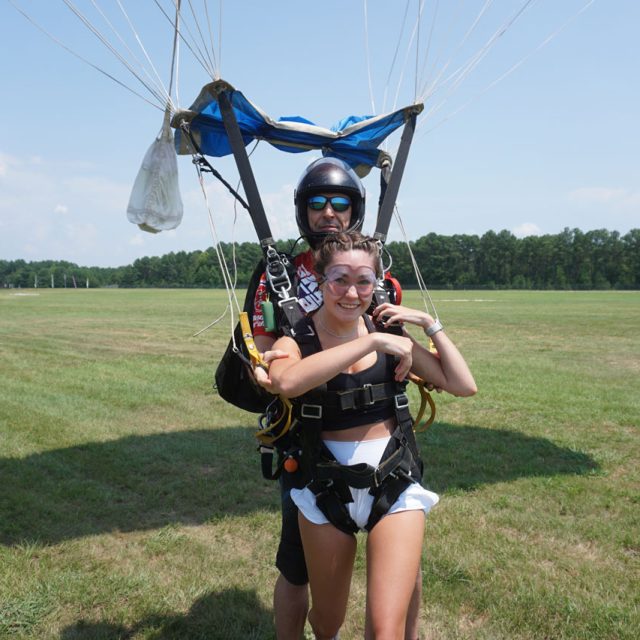 A tandem skydiving pair has just landed on a grassy field. The smiling woman in front is wearing a harness, and the instructor behind her is in full gear, including a helmet and sunglasses. The parachute is visible above them. Trees are in the background.