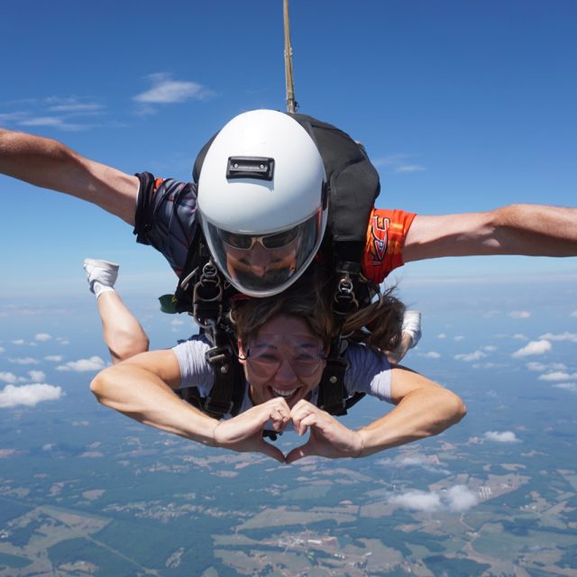 Two people skydiving tandem, with a bright blue sky and clouds in the background. The person in front is making a heart shape with their hands, while the instructor guides from behind. Both are wearing helmets and goggles.