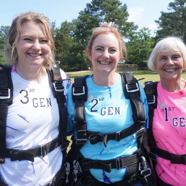 Three smiling women wearing skydiving harnesses stand outdoors. Their shirts read "3rd Gen," "2nd Gen," and "1st Gen," indicating different generations. Trees and a grassy area are visible in the background.