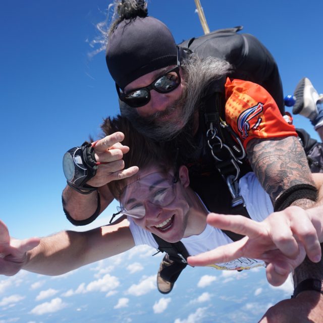 Two people tandem skydiving, smiling and making peace signs. The instructor has a black cap and sunglasses, while the other person wears goggles. The background shows a clear blue sky and clouds beneath them.