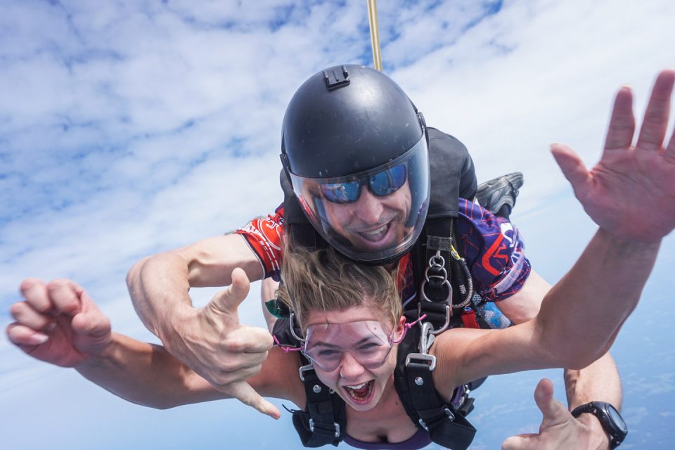 A woman and her instructor are skydiving tandem. They're free-falling through a clear sky with scattered clouds. Both appear excited, making gestures with their hands and smiling widely as they descend. The instructor wears a helmet and sunglasses.