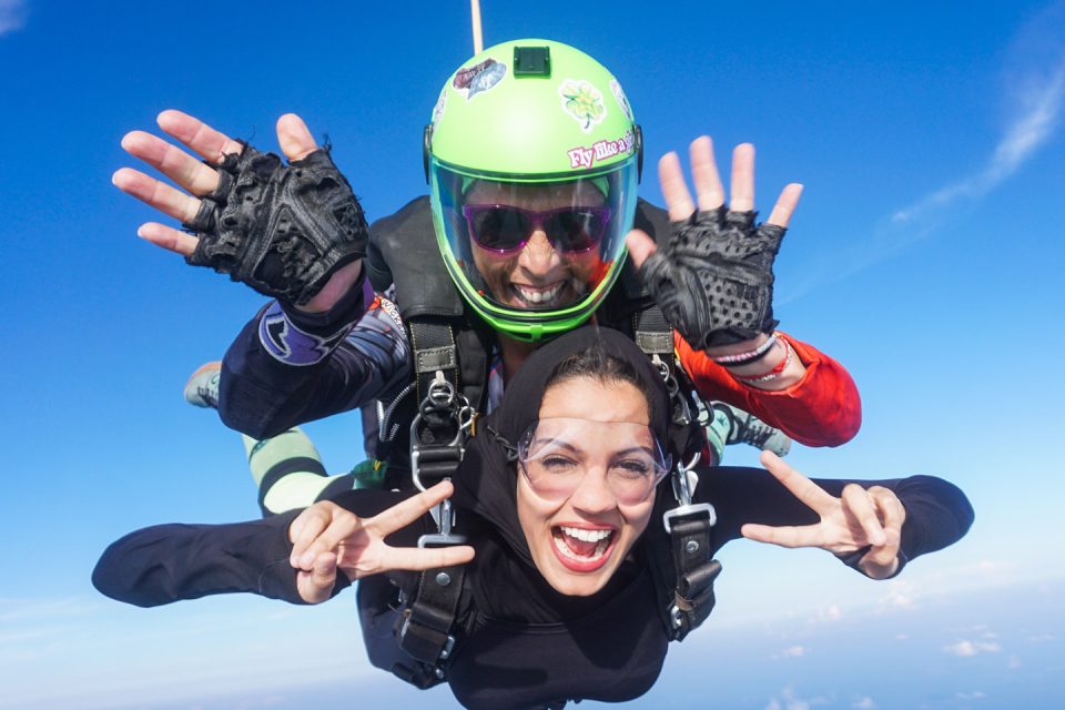 Two people tandem skydiving against a clear blue sky. The person in front is smiling widely and gesturing with peace signs, while the instructor behind is wearing a green helmet and waving with both hands. Both wear goggles and harnesses.