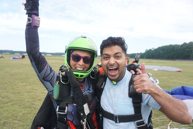 Two people celebrate after a tandem skydive. One is wearing a green helmet and parachute gear, and the other gives a thumbs-up. They stand on grassy ground, with parachutes visible in the background and a clear, blue sky overhead.