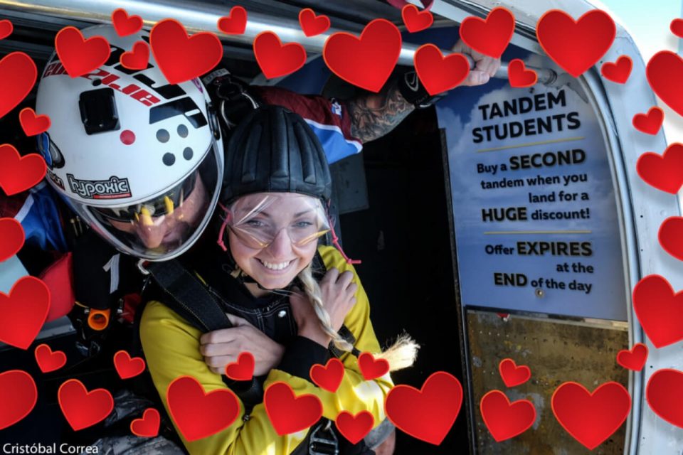 Two people in skydiving gear prepare to jump from an aircraft. Red heart graphics surround them. A sign in the background offers a tandem skydiving discount for students. They're smiling and appear excited for the adventure.