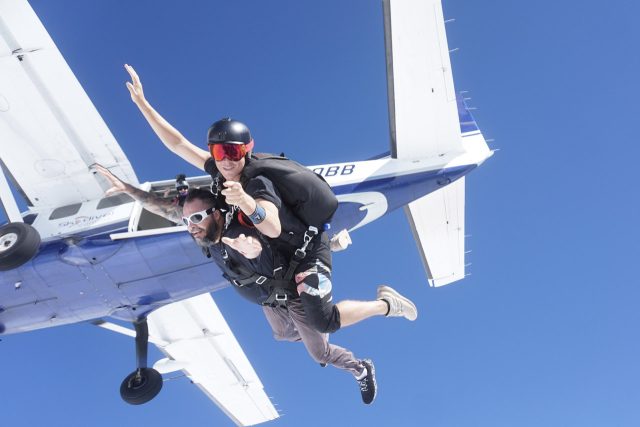 Two people are tandem skydiving, with one wearing a black helmet and goggles. They are free-falling next to a blue and white aircraft against a clear blue sky.