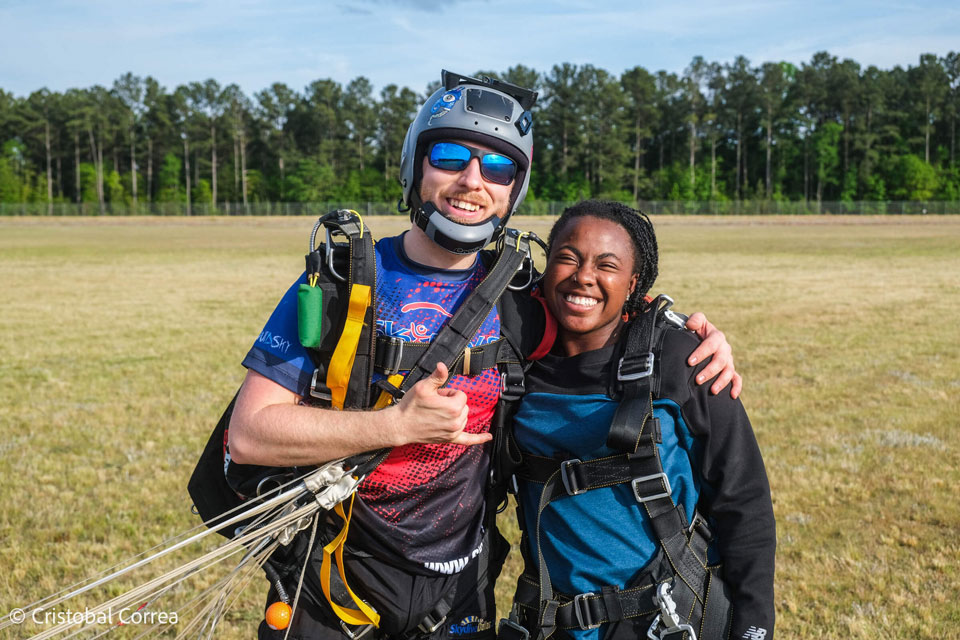 Two people wearing parachute gear stand smiling on a grassy field. One person gives a thumbs-up, while both appear happy and excited. Trees and a clear sky are in the background.