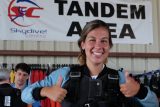 A person wearing a skydiving harness gives two thumbs up in a tandem area. There are skydiving signs and equipment in the background.