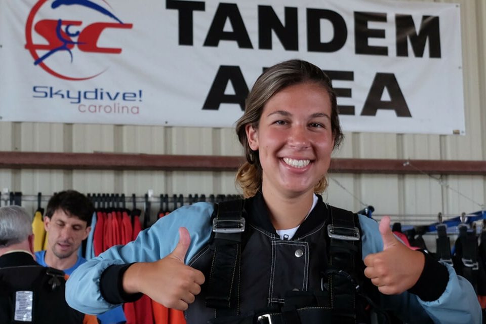 A person wearing a skydiving harness gives two thumbs up in a tandem area. There are skydiving signs and equipment in the background.