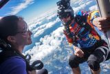 A woman and a man in colorful skydiving gear smile at each other inside a plane. The man holds the plane's door frame. They wear harnesses and goggles, ready to jump, with a scenic view of clouds and landscape below.