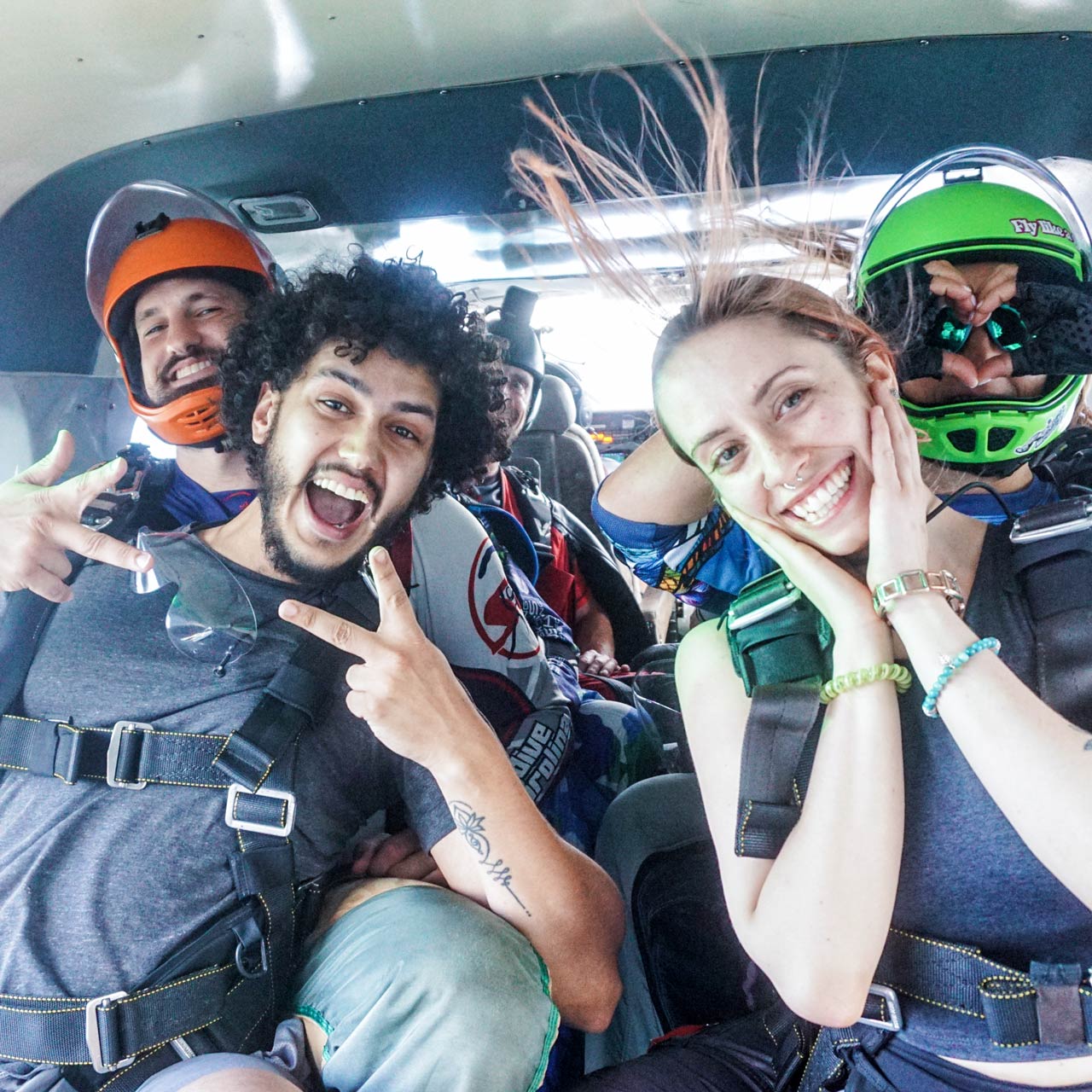 A group of skydivers inside an aircraft, smiling and showing excitement. One person in the front center gives a peace sign, while another beside them holds their face with hands. Two others in the background wear helmets and harnesses, ready for the jump.