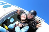 A woman in a blue jumpsuit and helmet smiles excitedly as she prepares to skydive with an instructor beside her. They are both at the open door of an airplane, ready to jump, against a clear blue sky backdrop.