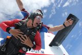 Two people skydiving, tandem jumping from an airplane. The front person looks excited with arms wide, wearing a black and red jumpsuit and goggles. The sky is clear with a few clouds, and the plane is partially visible in the background.